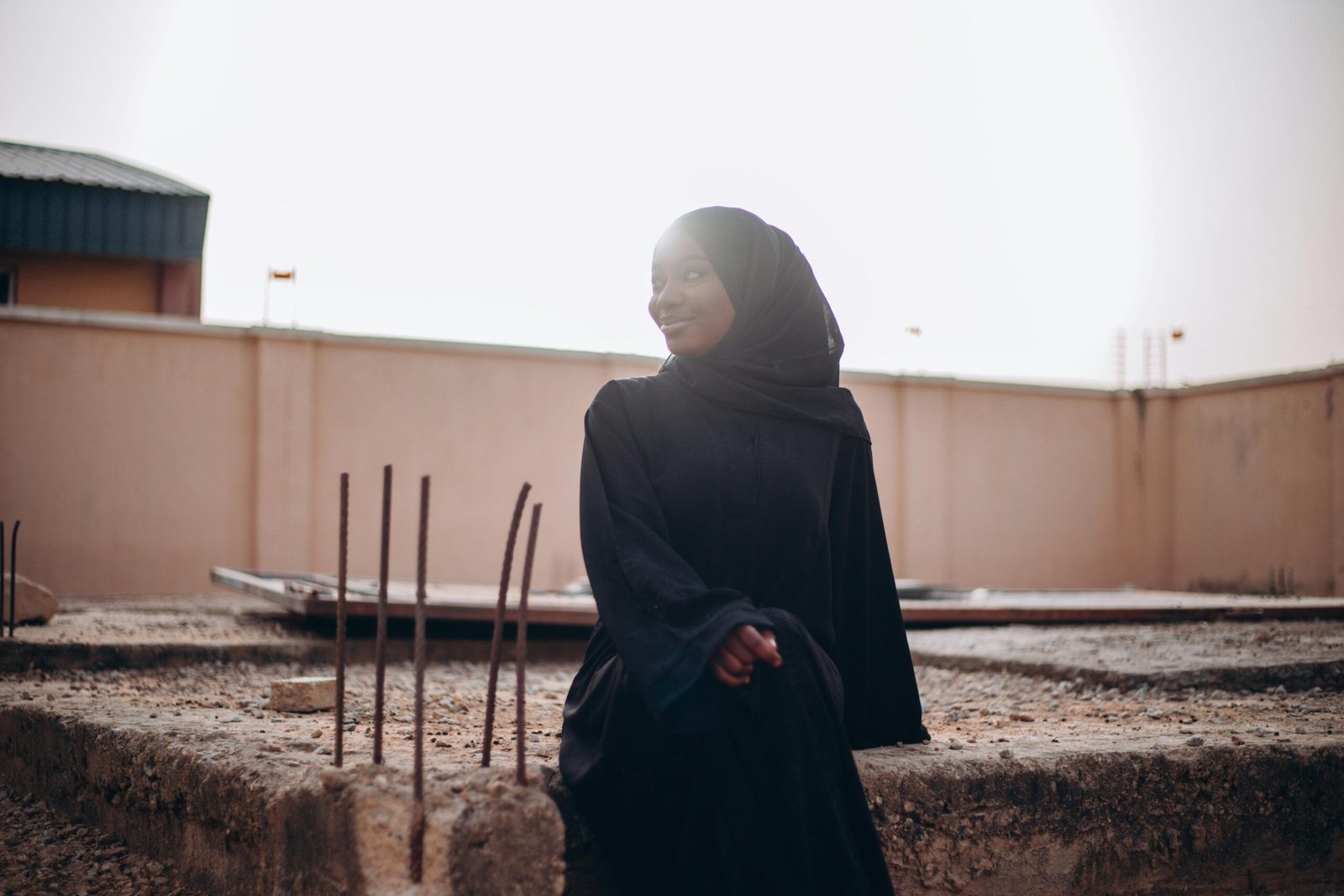 A young woman in a black hijab sits at a construction site during sunset, exuding a serene mood.